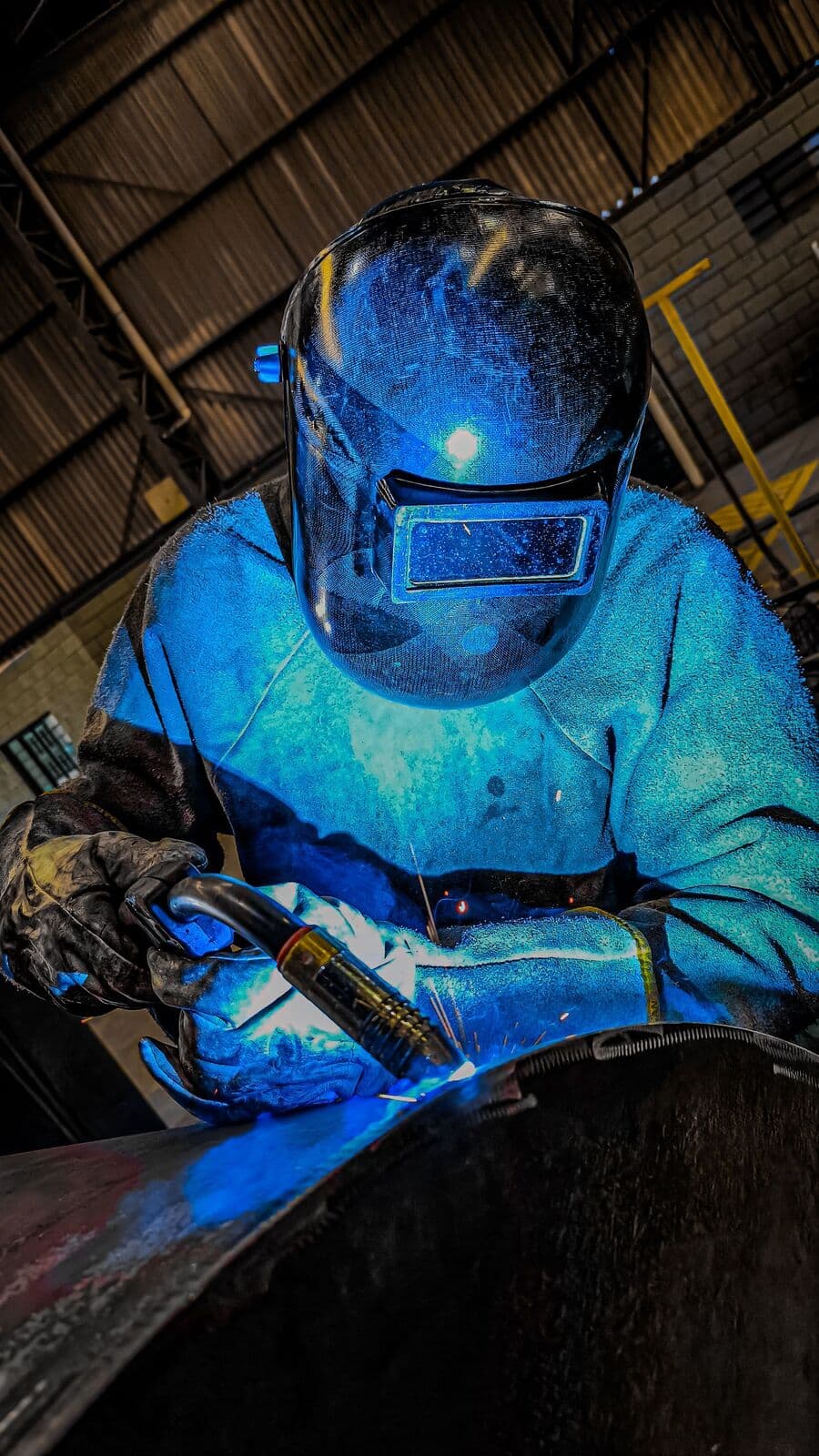 Soudeur au masque penché sur une pièce, halo bleu de l'arc dans l'atelier