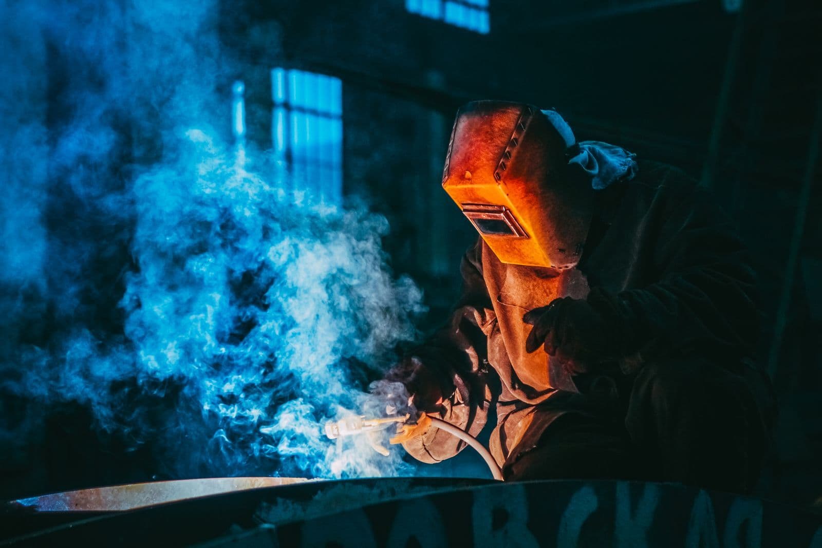 Soudeur au masque en train de souder une pièce, fumée bleue dans l'atelier sombre